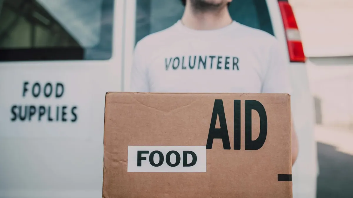 Smiling volunteer holding a box labeled 'Food Aid' outside a van with 'Food Supplies' sign.