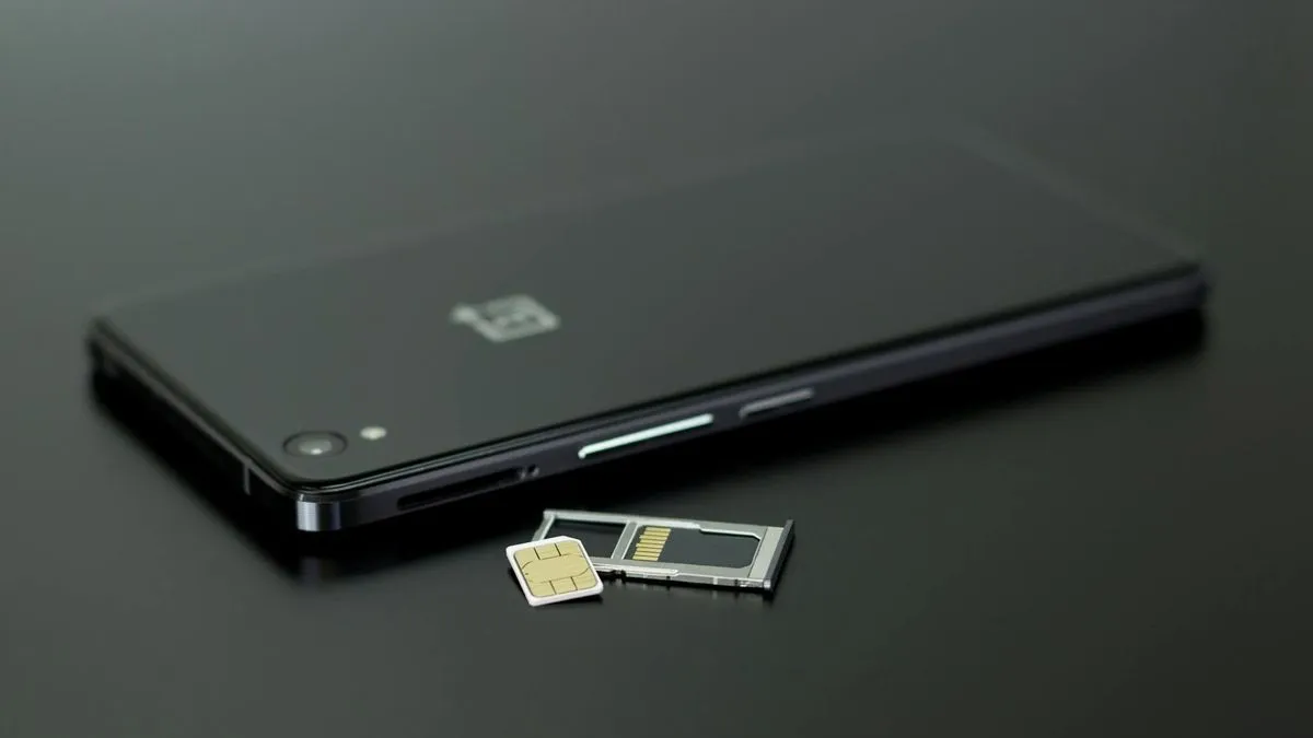 A black smartphone lies on a dark surface next to a SIM card and its tray.
