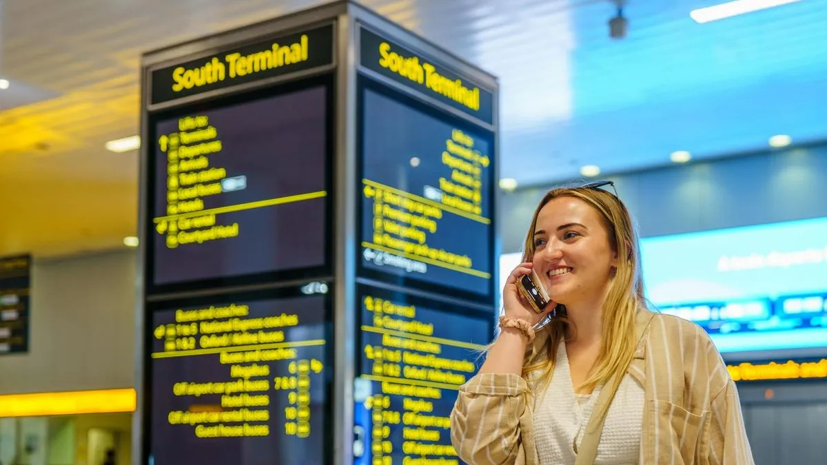 A woman smiles while talking on her phone in an airport terminal next to a departure board.