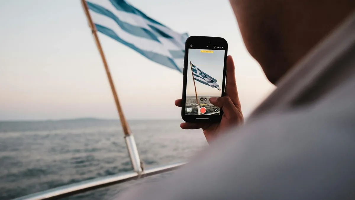 A person captures the Greek flag on a smartphone while sailing on a calm sea during the day.