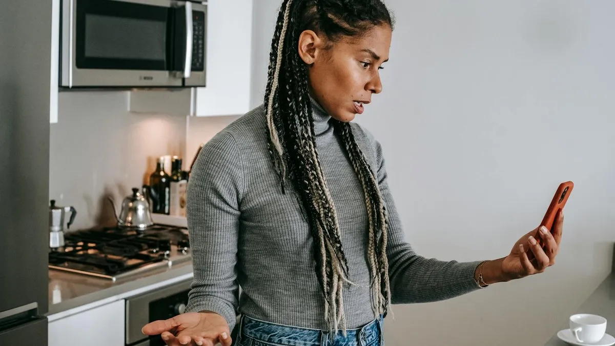 A woman with braided hair looks confused and frustrated while holding her phone in a kitchen.