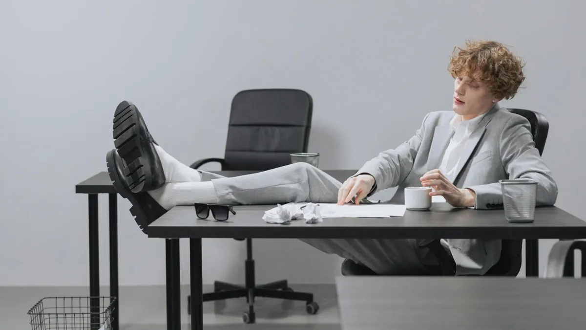 A curly-haired man in a suit relaxes with feet on desk, looking serious in a modern office setting.