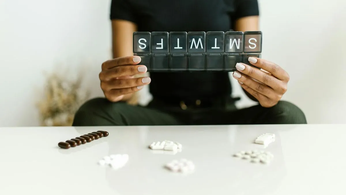 A person holding a weekly pill organizer, with various pills scattered on a white table.