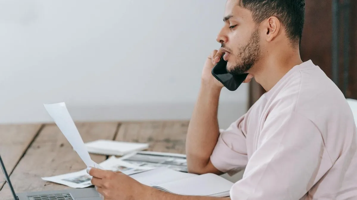 A man on the phone, looking at documents and a laptop, appearing to discuss a contract.