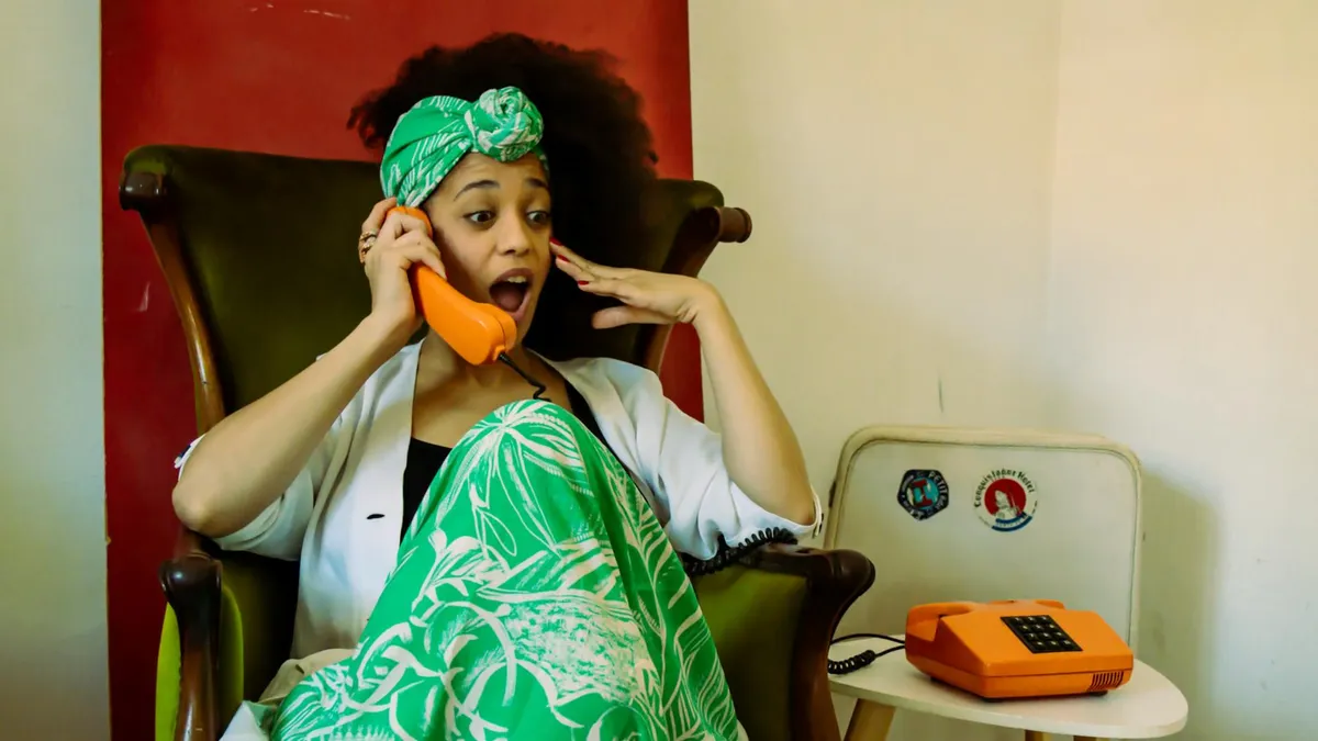A woman in vibrant attire is surprised while talking on a retro orange telephone indoors.