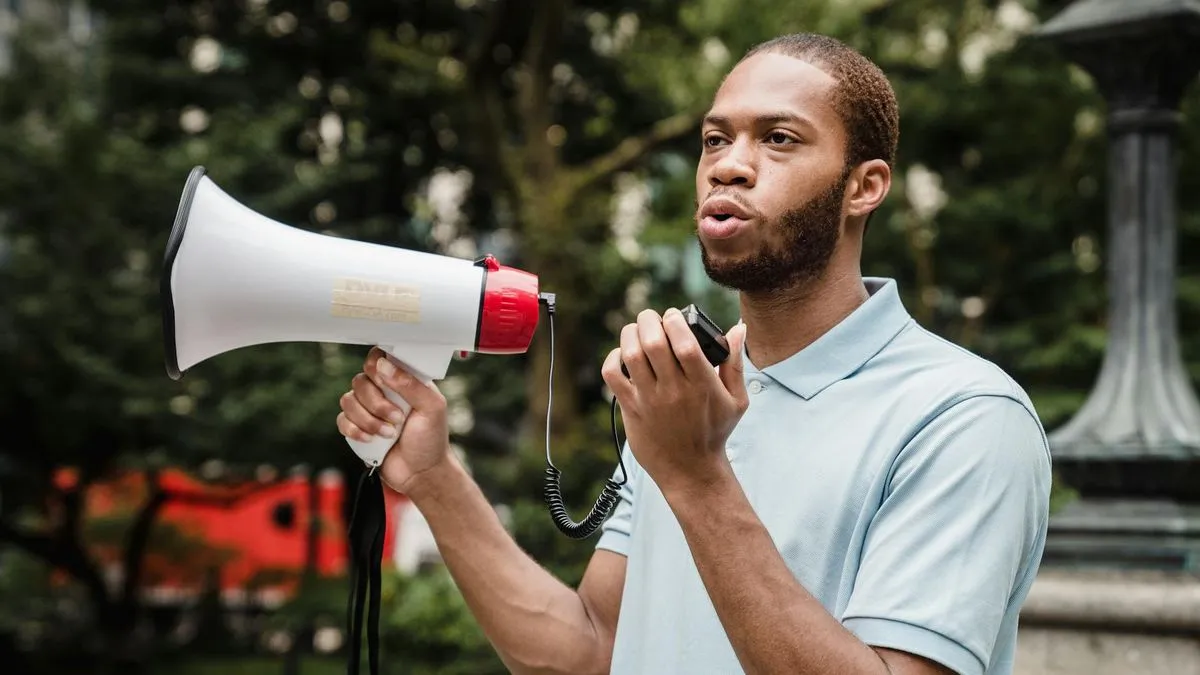 A man in a light blue polo shirt speaks into a white and red megaphone outdoors.