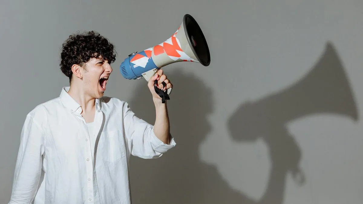 A person with curly hair shouts into a megaphone, casting a large shadow on the wall.