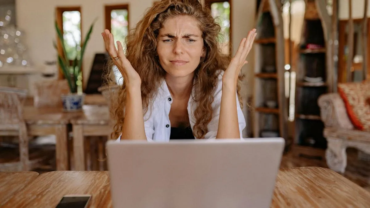 A frustrated woman with curly hair throws her hands up in confusion while looking at a laptop.