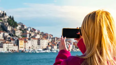 A woman takes a photo of the Šibenik waterfront using a smartphone on a sunny day.