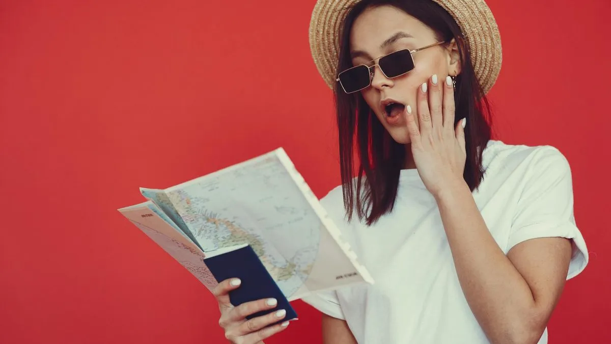 A surprised woman in a straw hat and sunglasses holds a passport and a map against a red background.