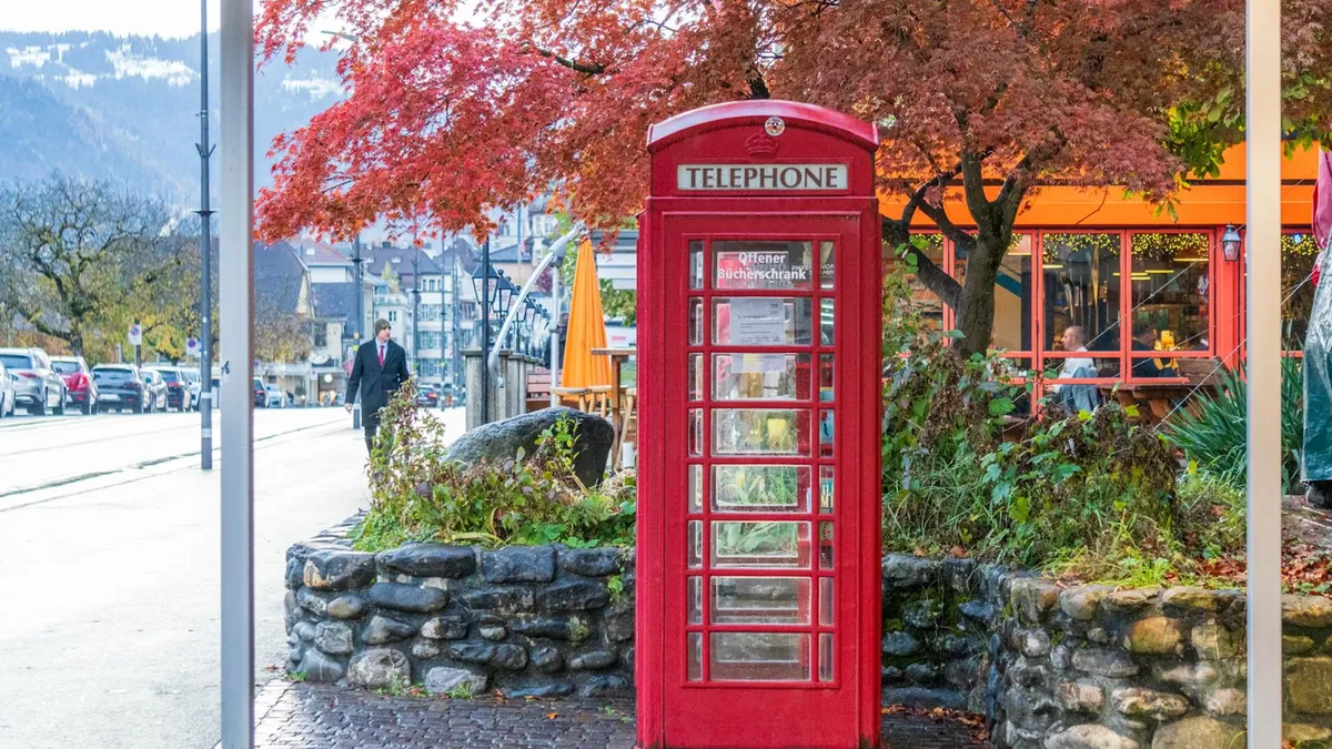 Red telephone booth amidst vibrant autumn foliage in Interlaken, Switzerland street scene.