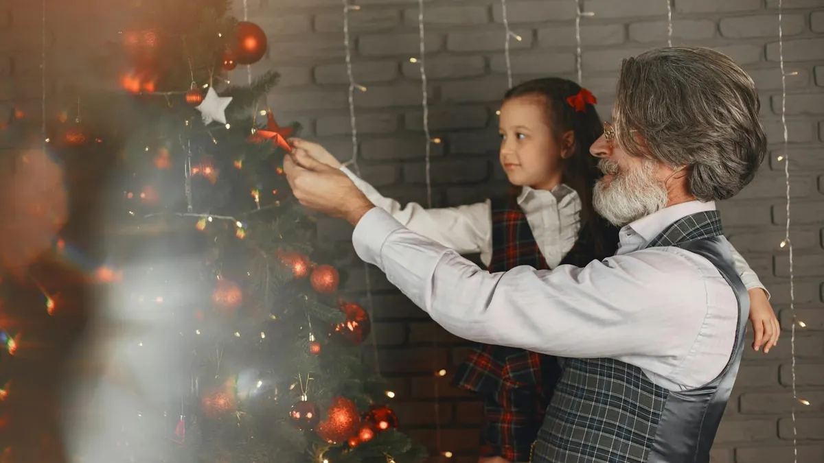 A grandfather helps his granddaughter place an ornament on a Christmas tree, capturing a moment of holiday togetherness.