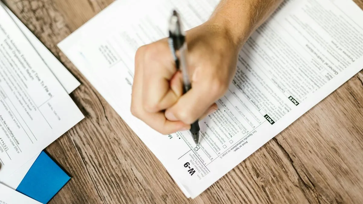 A person's hand holding a pen, filling out a form on a wooden desk.