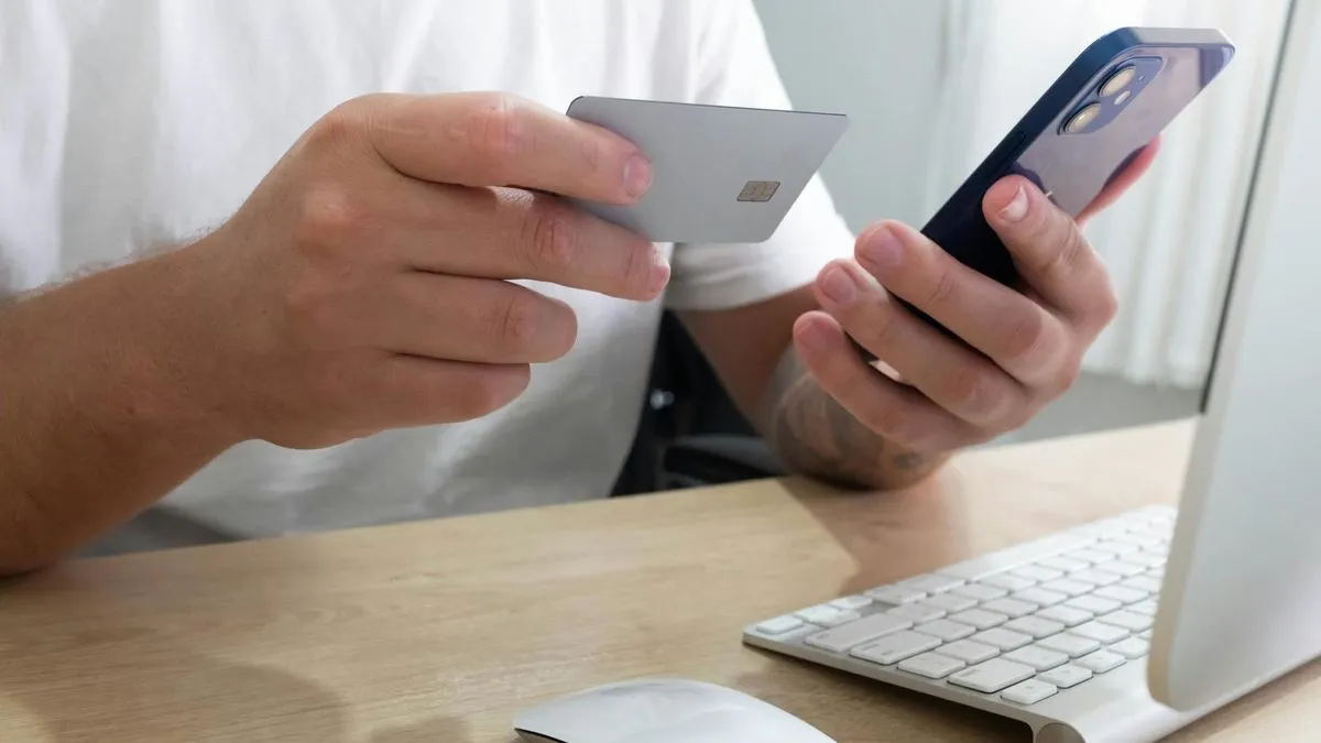 A person holds a credit card and a smartphone, with a computer keyboard and mouse on a desk.