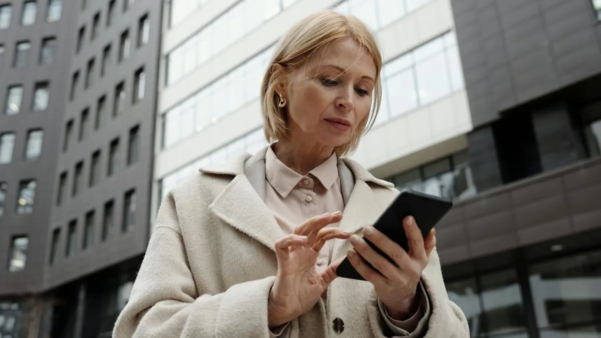 A woman in a beige coat looks intently at her smartphone while standing outside a modern building.