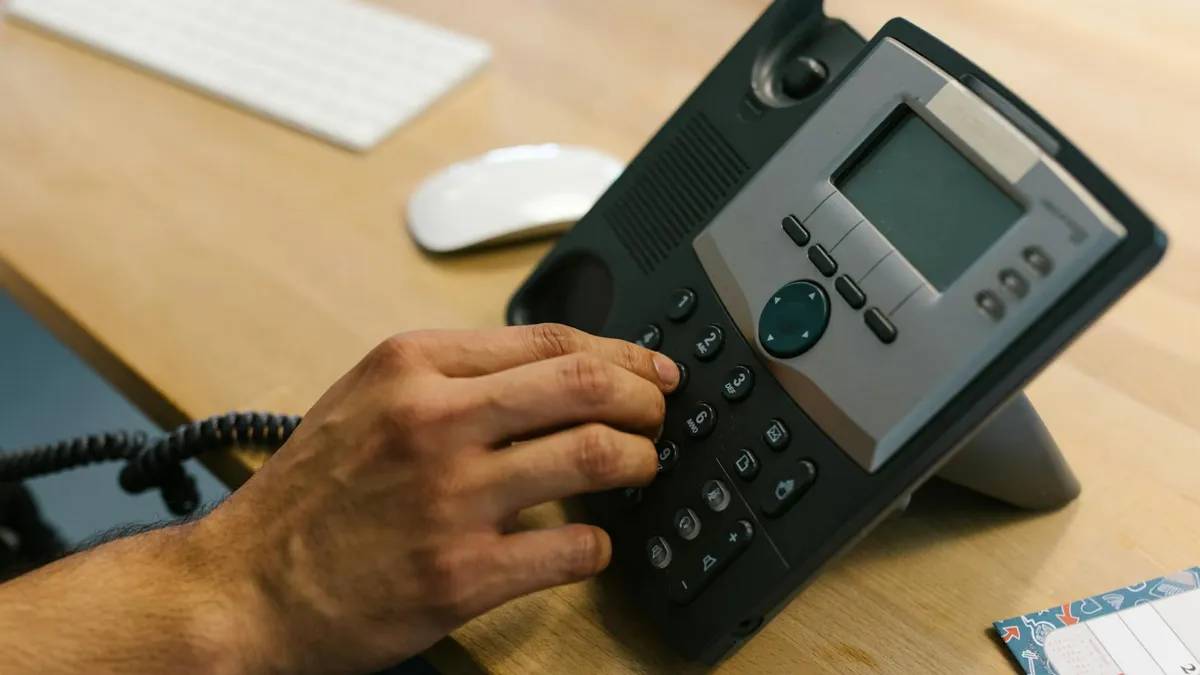 A hand dialing a phone on a wooden desk. Office environment with keyboard and planner.