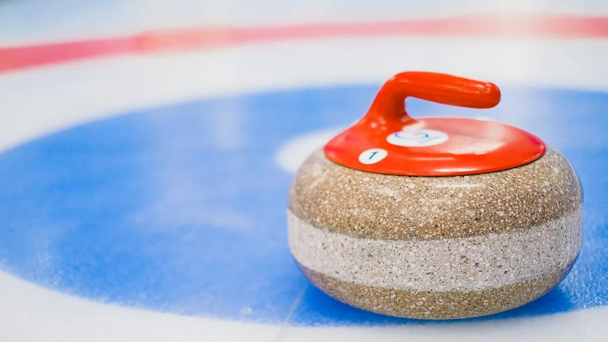 A red-handled curling stone rests on a blue and white target on an ice rink.