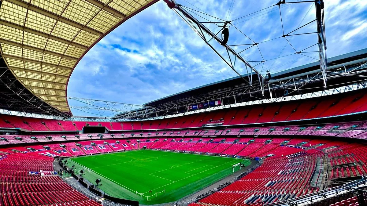 An empty Wembley Stadium with vibrant green pitch and red seats under a bright blue sky.