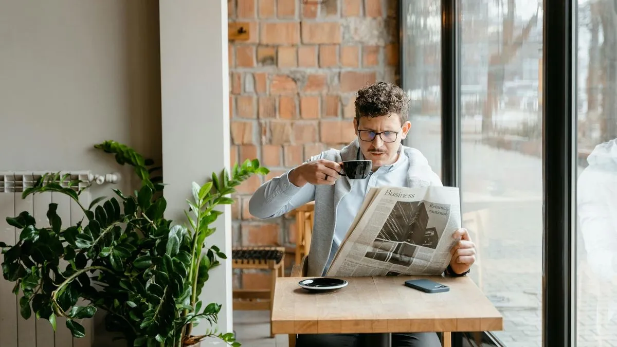 A man in glasses sips coffee while reading a newspaper at a wooden table in a cafe.
