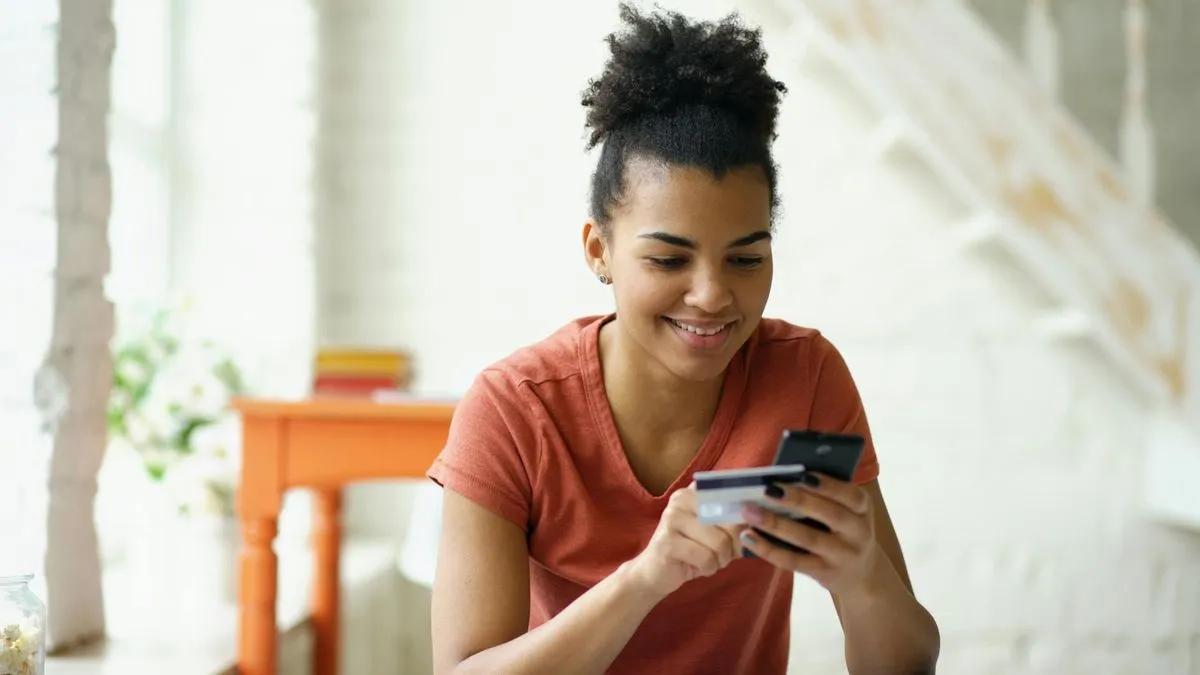 A smiling woman holds a smartphone and a credit card, appearing to make an online payment.
