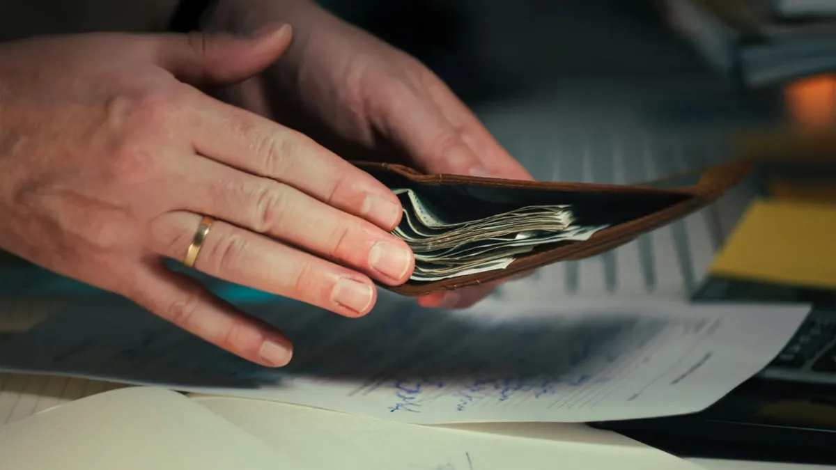 Hands holding an open wallet filled with cash above documents, indicating financial planning or budgeting.