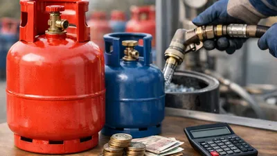 A red and a blue gas bottle on a wooden surface, with coins, banknotes, and a calculator. A gloved hand holds a nozzle refilling a gas bottle.