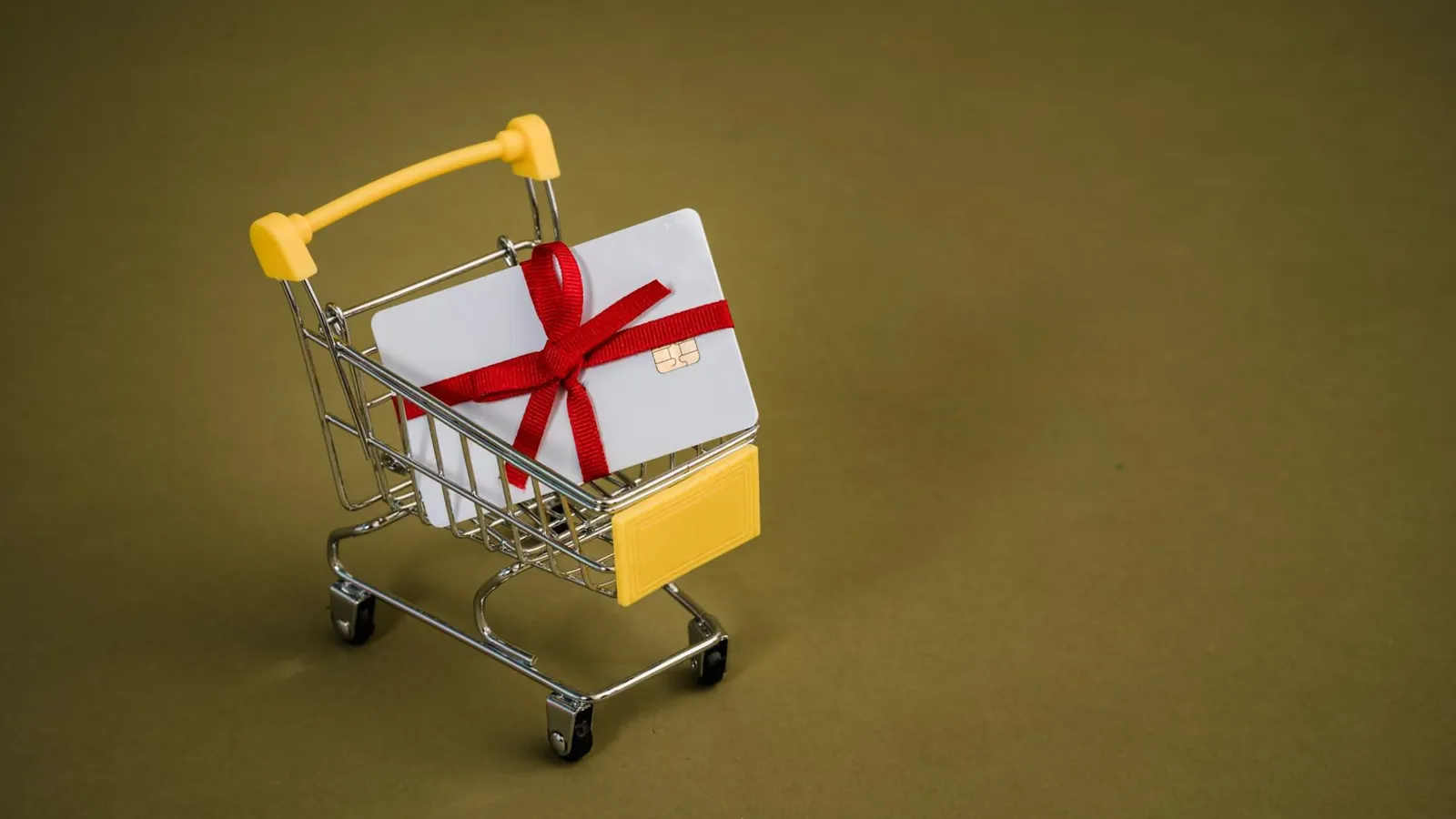 A gift card with a red bow sits in a mini shopping cart on a yellow backdrop.