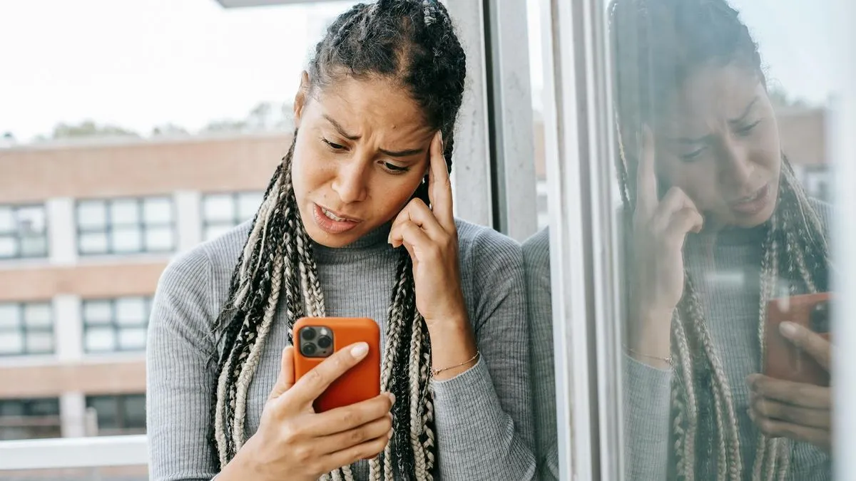 A woman with braided hair looks frustrated while holding and staring at her orange smartphone.