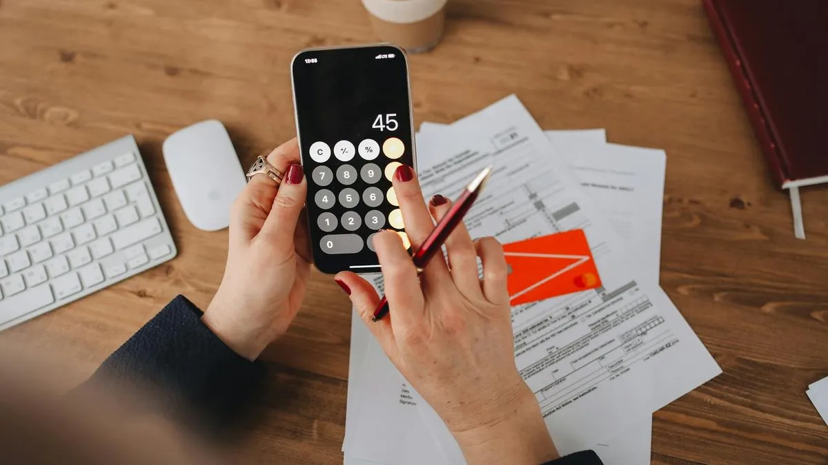 A person's hands using a smartphone calculator with bills and a credit card on a wooden desk.