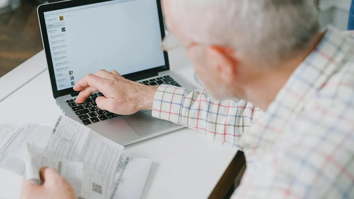 An older man in a plaid shirt uses a laptop while holding a document.