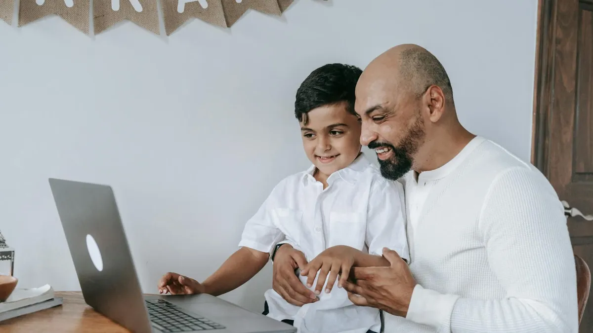 A father and son smiling while using a laptop, creating a moment of joy and bonding indoors.