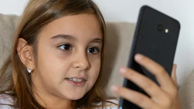 A young girl with long brown hair smiles while using a smartphone indoors.