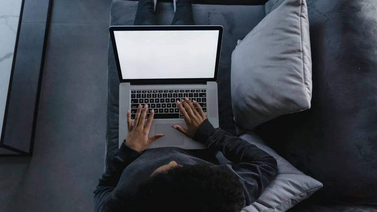 Overhead shot of a person lying on a grey sofa, typing on a laptop with a blank screen.