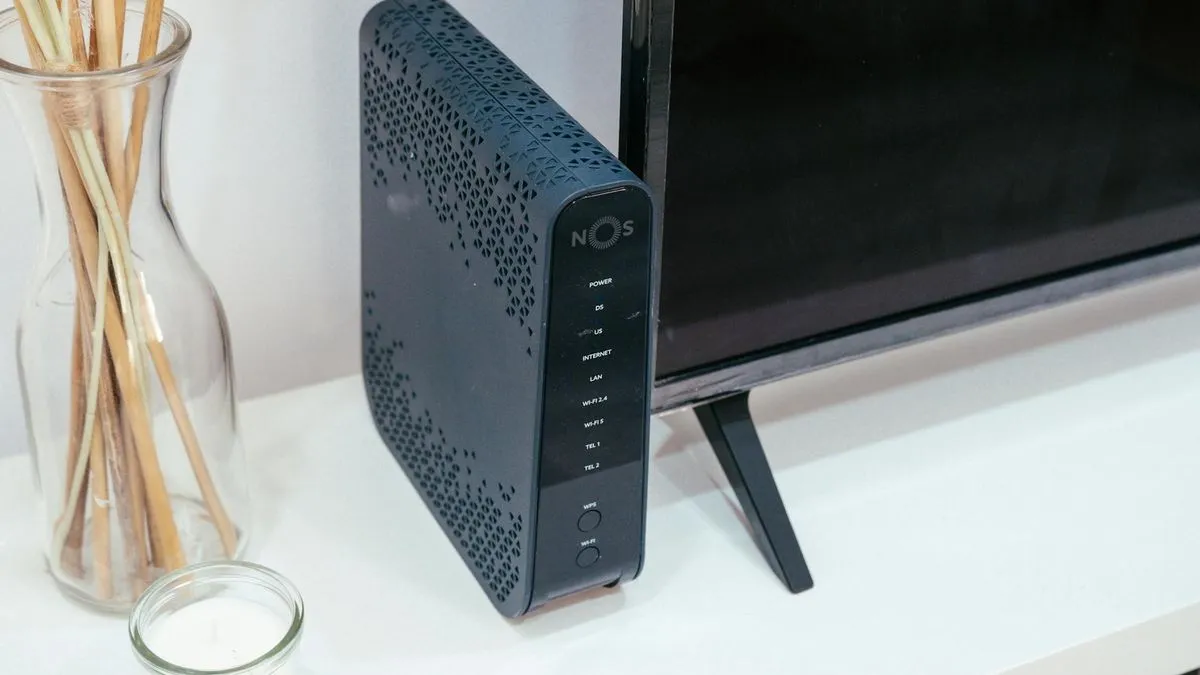 A dark grey internet router with a NOS logo stands upright next to a television.