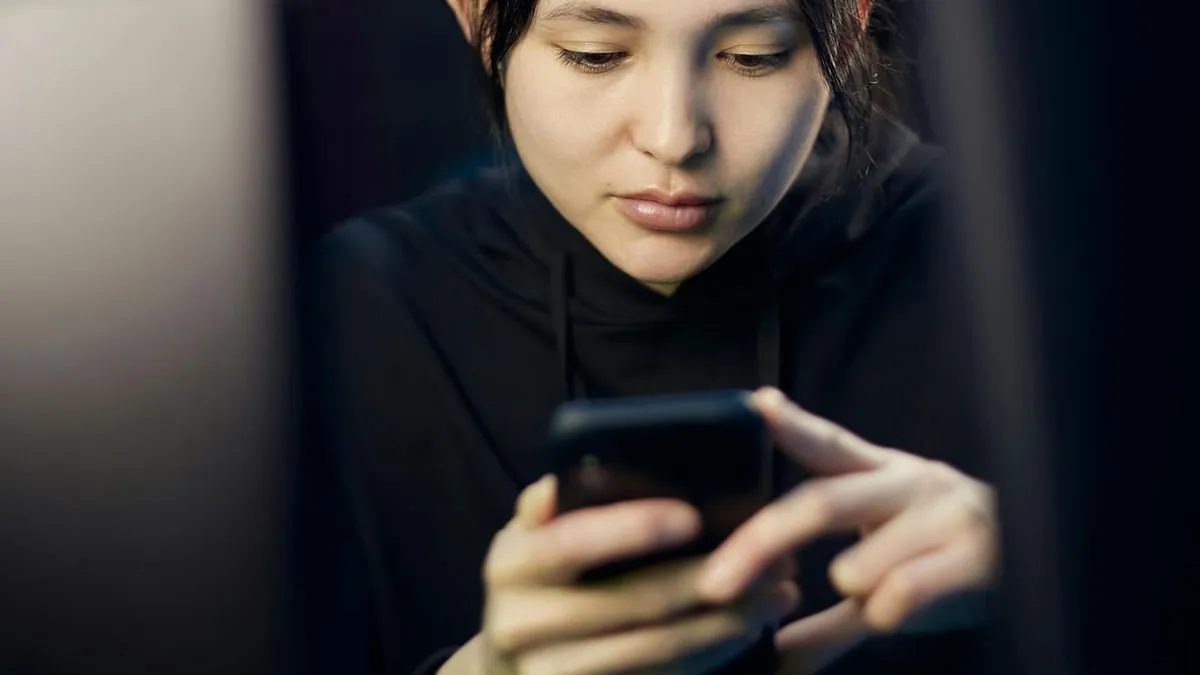 A young woman in a dark room intently looking at and interacting with her smartphone.