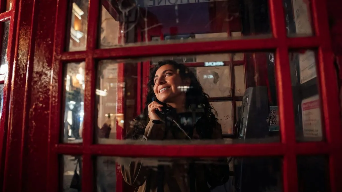 A smiling woman with dark curly hair talks on a phone inside a red British telephone booth.