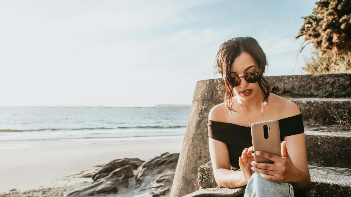 A woman sitting on beach stairs looking at a smartphone, enjoying a sunny day by the sea.