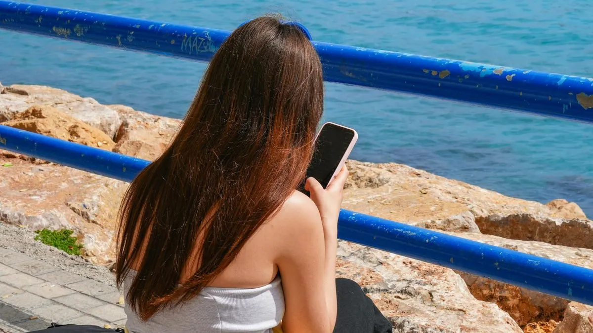 A woman with long brown hair sits by the sea, looking at her phone.