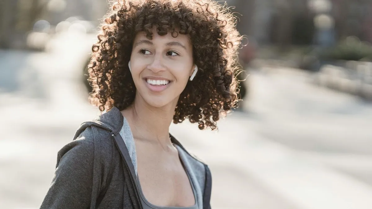 A smiling woman with curly hair wearing white earbuds, looking to the side.