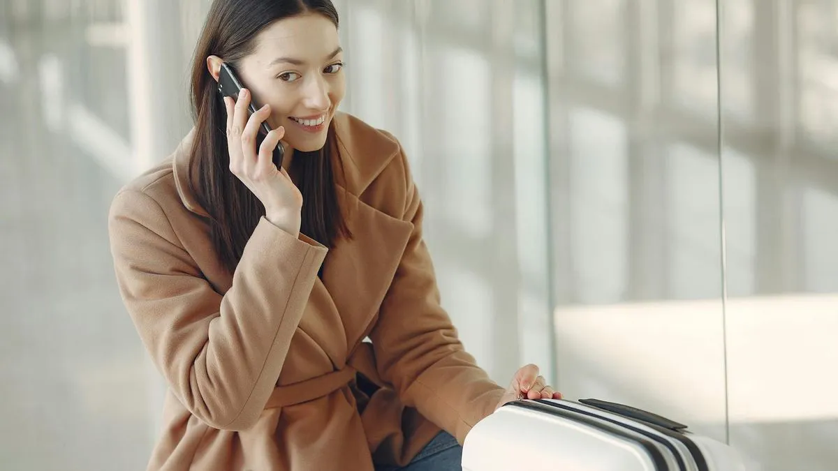 A woman in a brown coat talks on a phone next to a white suitcase.