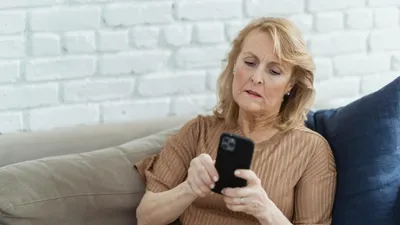 A woman with blonde hair sits on a sofa, looking intently at her black smartphone.