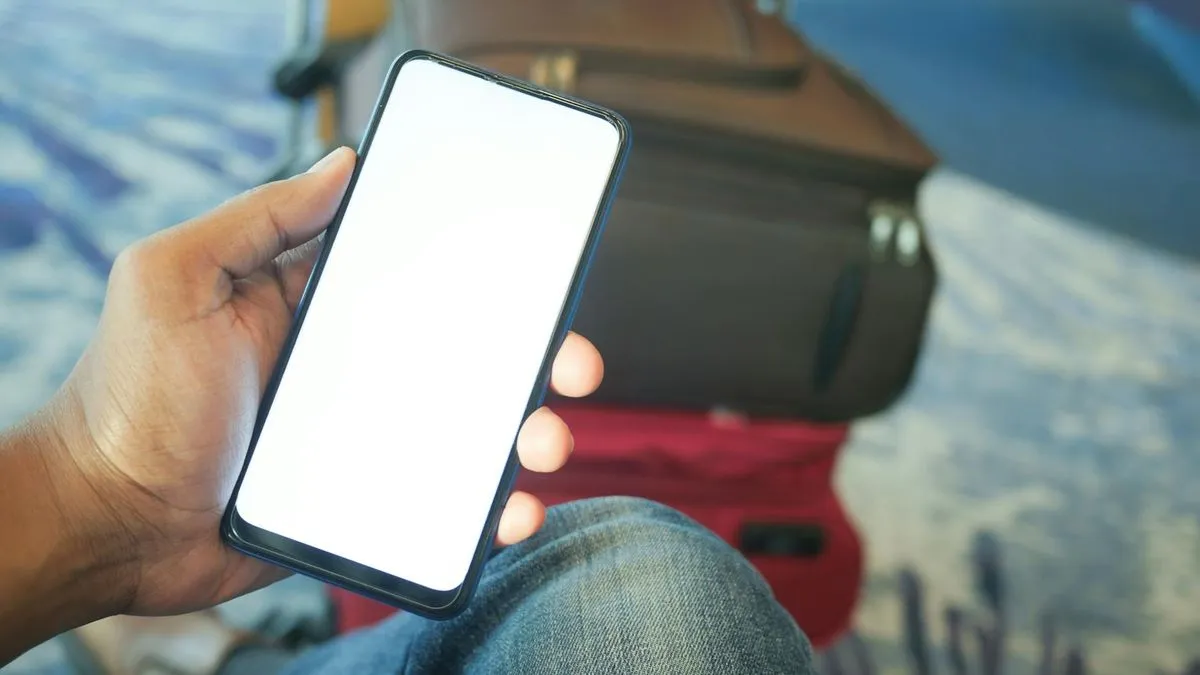 A person holds a smartphone with a blank white screen, with luggage visible in the background.