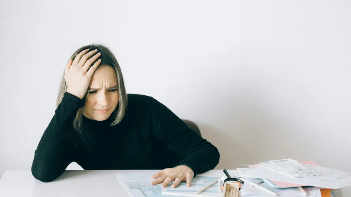 Woman in black sweater stressed with financial paperwork, overwhelmed at white table.