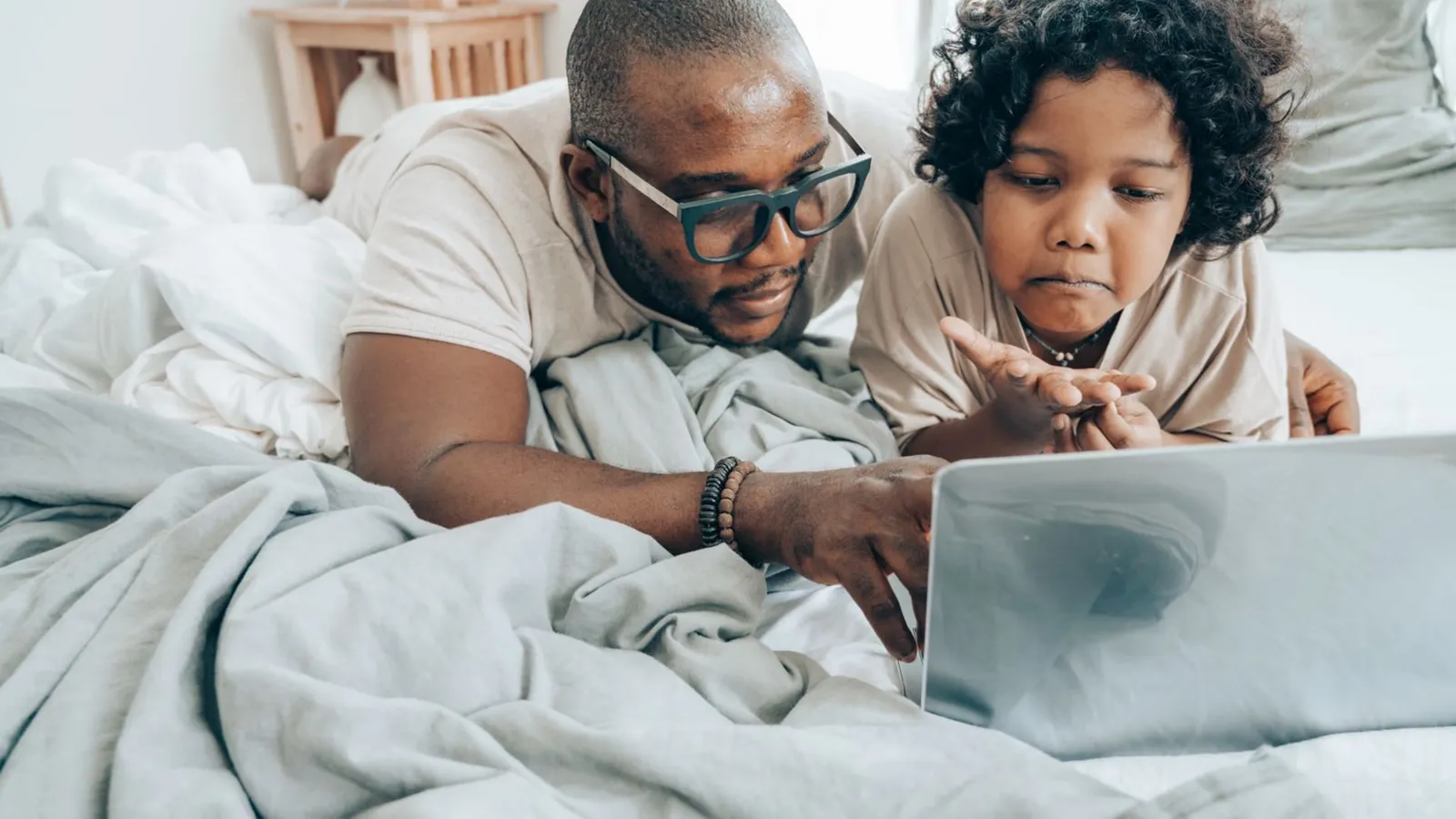 Focused African American male in glasses and child with curly hair in pajama lying on bed in bedroom and using laptop