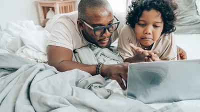 Focused African American male in glasses and child with curly hair in pajama lying on bed in bedroom and using laptop