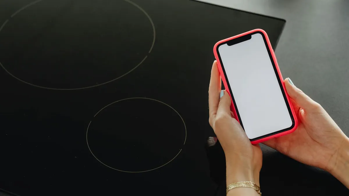 Close-up of hands holding a smartphone with a blank screen, beside a black cooktop.