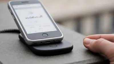 A silver smartphone with 'vodafone' on its screen, resting on a black wireless charger. A hand is nearby.