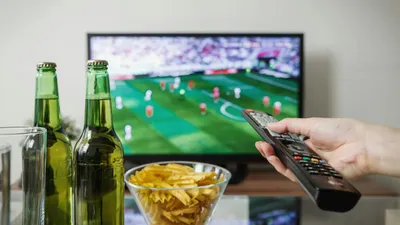 A hand holding a TV remote control, with a football match on a TV screen in the background.