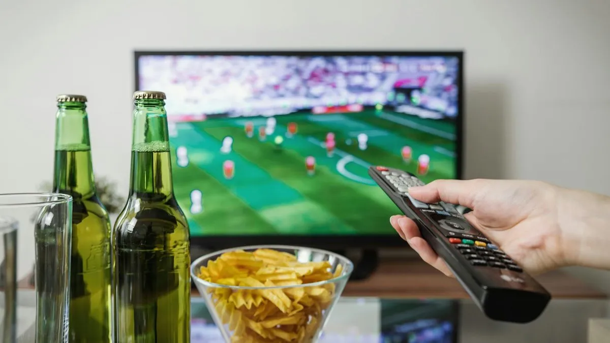 A hand holding a TV remote control, with a football match on a TV screen in the background.