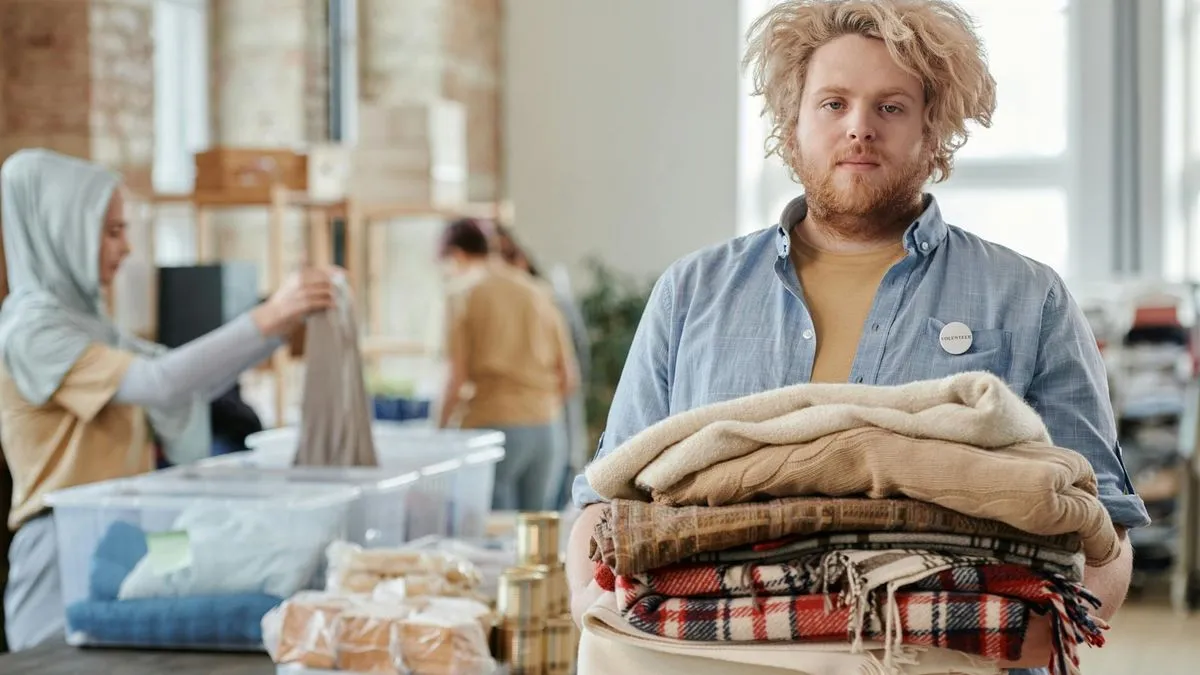 A man with curly hair holds a stack of folded clothes, with other people sorting items in the background.
